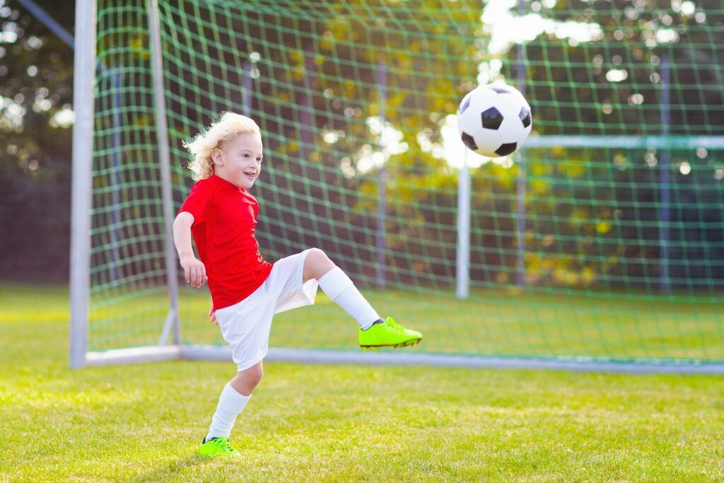 Kids play football on outdoor stadium field. Children score a goal during soccer game. Little boy kicking ball.